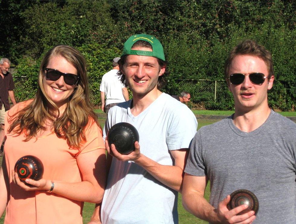 Two men and a woman holding lawn bowling balls on the Central Park greens with players in the background