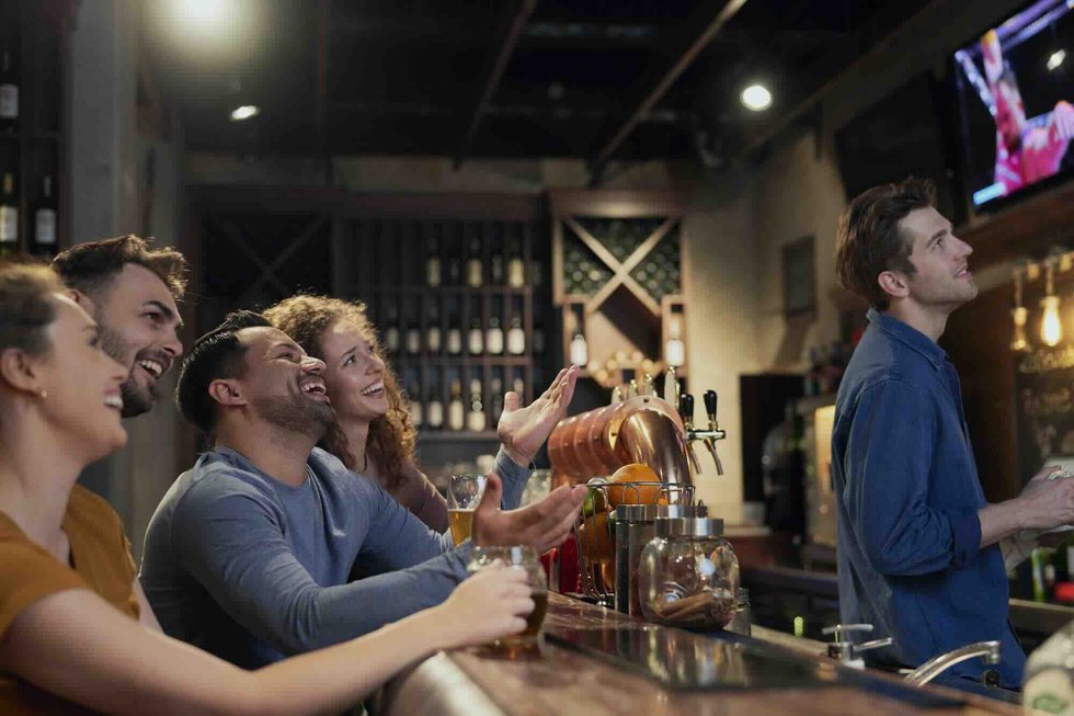 Two men and two women laughing with beers at a bar, with a bartender and beer taps in the background.